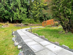 A pathway with stone slabs and gravel, surrounded by grass and trees, leading to a wooden structure. - Lavender Suite on the Dungeness River (Sequim)