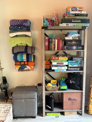 Ein Zimmer mit einem Bücherregal voller Brettspiele, einem grauen Hocker und einem an der Wand befestigten Regal mit gefalteten Decken.