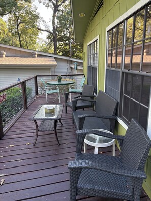 Eine überdachte Terrasse im Freien mit Rattanmöbeln und einem Glastisch, umgeben von einem grünen Gebäude mit großen Fenstern.