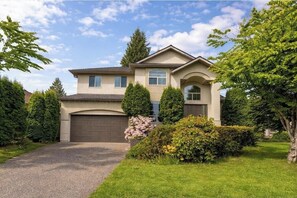 A two-story house with a garage, surrounded by well-maintained lawns and trees. - Surrey-Langley-Luxurious Suite | Prime Location & Ultimate Comfort (Langley)