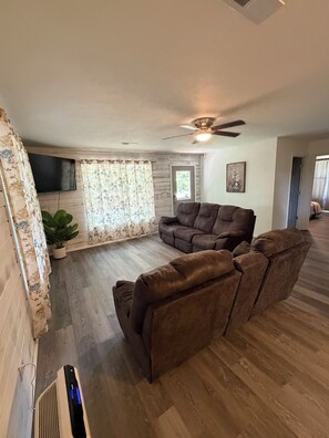 A living room with a sectional sofa, a ceiling fan, and a flat-screen TV mounted on the wall.