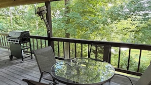 Een overdekt terras buiten met een glazen tafel, metalen stoelen, een barbecue en uitzicht op een bebost gebied.
