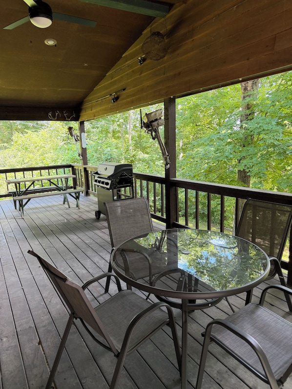 A covered outdoor deck with a round glass table and metal chairs, a grill, and a view of a wooded area.