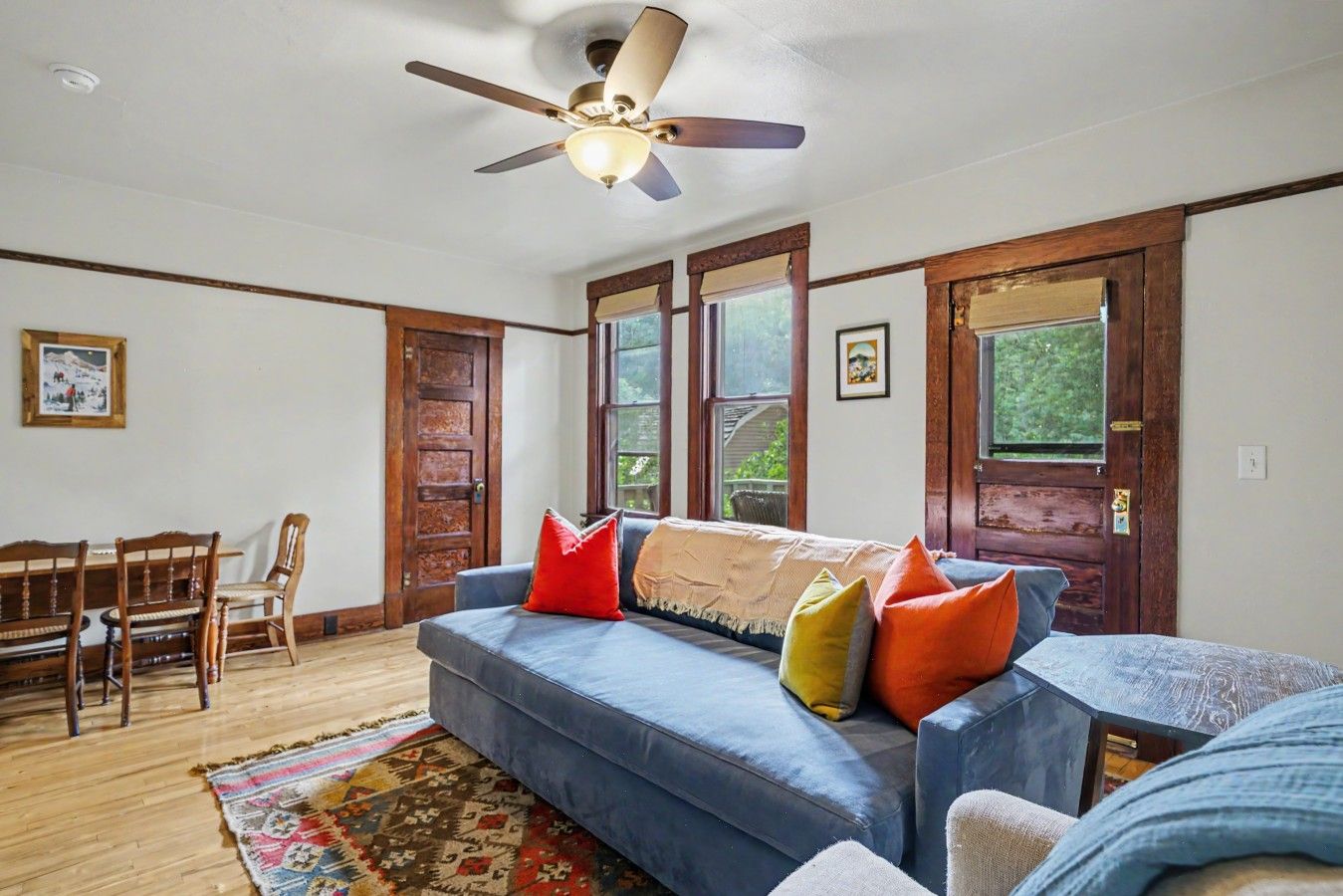 A living room with a blue sofa, wooden chairs, a ceiling fan, and a large window.