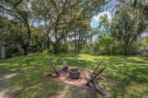 An outdoor area with a fire pit surrounded by wooden logs, a grassy lawn, and trees. - Riverfront Retreat | Sleeps 14 | Pool | Spa | Deck (Tampa)