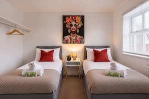 A hotel room with two beds, each with a red pillow and a towel. There is a small table with a lamp and a plant. A painting is on the wall above the beds. - Oriel House • Sleeps 6 • Free Parking • Work Desk • Fast WIFI • Self-Check-In • By Yoko Property (Milton Keynes)
