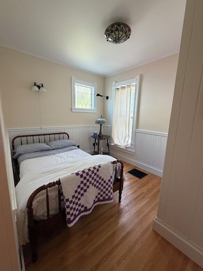 Ein Schlafzimmer mit einem hölzernen Bett, einer karierten Decke, einem Nachttisch, einer Lampe, einem Fenster mit Vorhängen und einer Deckenleuchte.