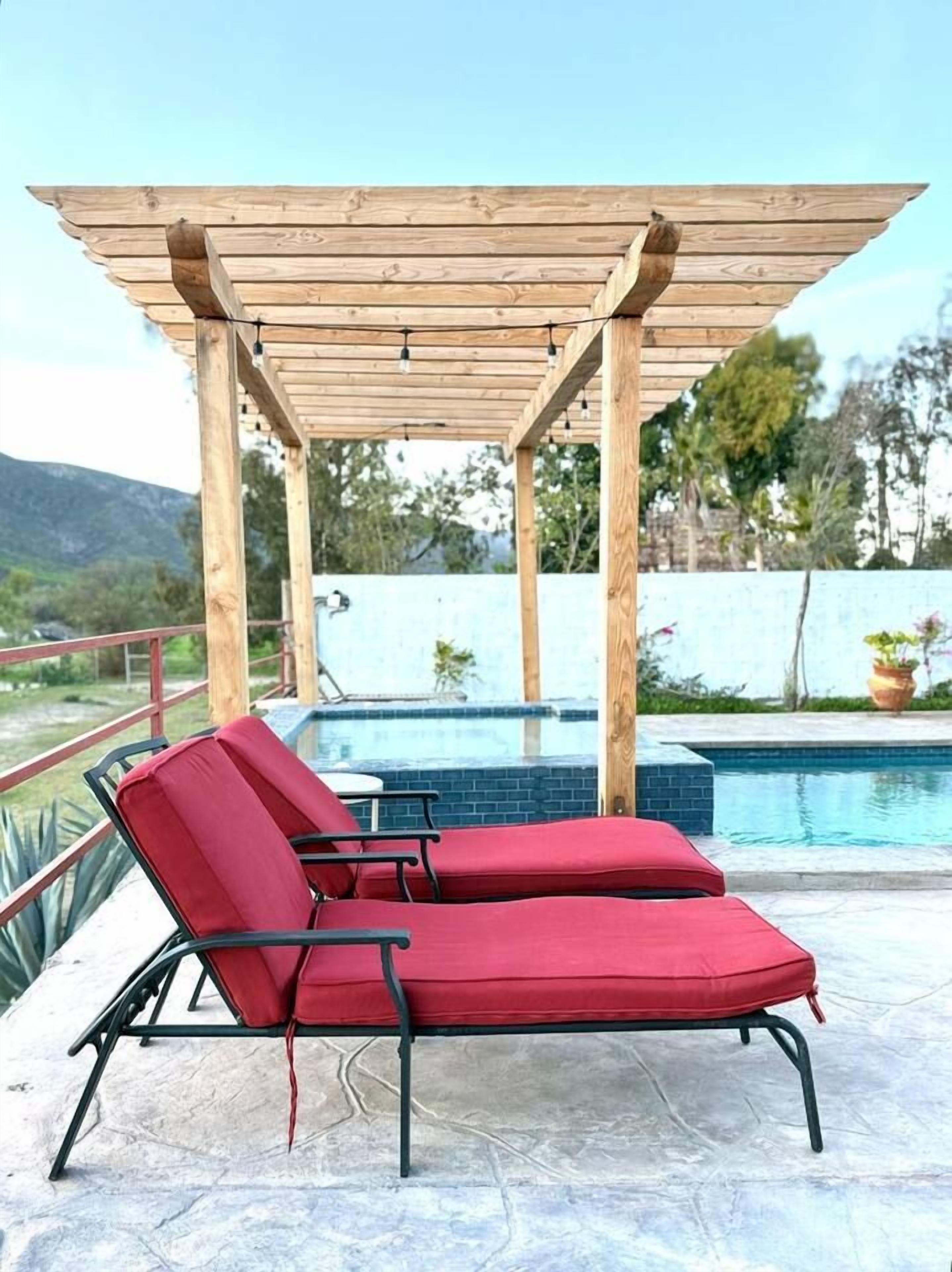Uma área na beira da piscina com duas espreguiçadeiras vermelhas sob uma pérgola de madeira, piscina e vista para a paisagem.