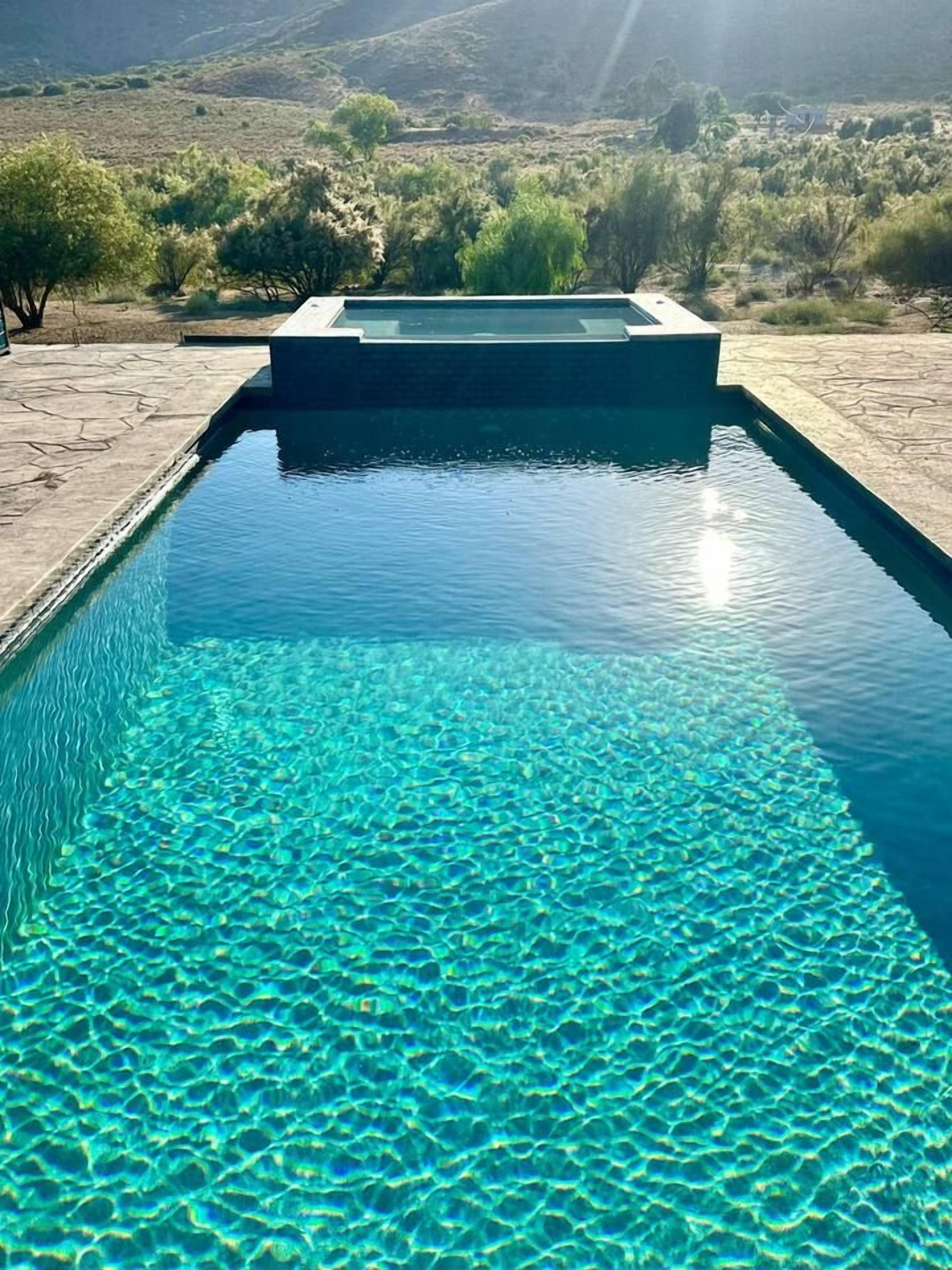 Une piscine rectangulaire à l’eau d’un bleu limpide, bordée de pierres, et entourée d’un espace aménagé avec une végétation clairsemée.