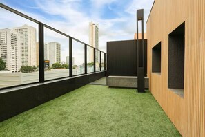 A modern interior with a view of tall buildings, a green artificial turf floor, and a wooden wall with a built-in bench. - Premium Apartment in Setor Bueno | Full-Service Condominium with Heated Pools (Goiânia)