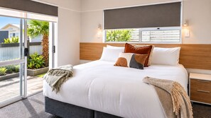 A modern bedroom with a large bed, a window with blinds, a bedside table, and a view of greenery outside. - Love It on Lemon | Stay Central | Explore More! (New Plymouth)