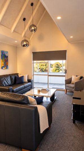 A modern living room with a sofa, coffee table, and armchairs, a painting on the wall, and a large window with a view. - Love It on Lemon | Stay Central | Explore More! (New Plymouth)