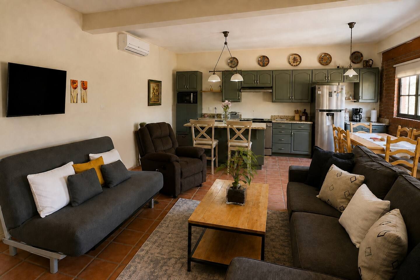 A living room with a sofa, armchairs, a coffee table, and a TV. There is a kitchen area with cabinets, a refrigerator, and a dining table.