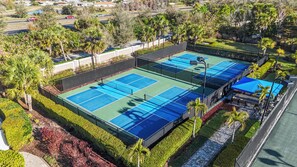Vue aérienne d’un complexe de courts de tennis comprenant plusieurs terrains, entouré d’espaces aménagés et de palmiers.