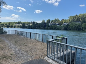 Un paseo fluvial con barandilla de metal, pavimento de piedra y vistas a casas al otro lado del agua.