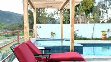 Área da piscina com duas espreguiçadeiras vermelhas sob uma pérgola de madeira, uma piscina e vista para a paisagem ao redor.