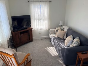 A living room with a television on a wooden stand, a grey sofa with patterned cushions, a wooden chair, and a small table with a remote control. - Quaint 2-bedroom bungalow, close to the ocean in charming Thomaston (Thomaston)