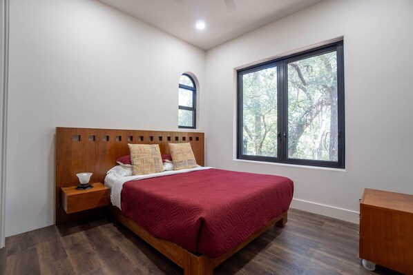 Un dormitorio con cabecera de madera, un ventanal con vistas a árboles, una cama con colcha roja y una mesita de noche con lámpara.