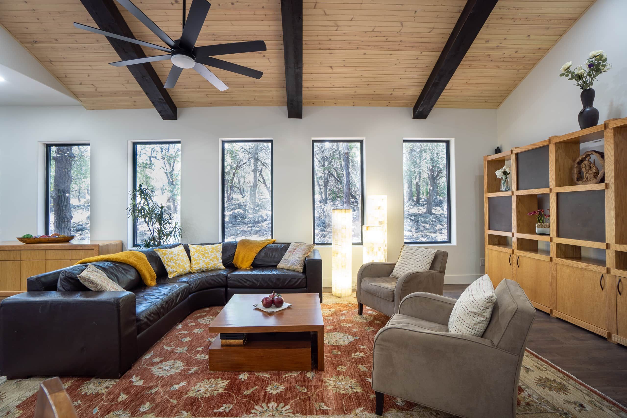 A modern living room with a sectional sofa, a coffee table, and a fireplace. There are large windows with a view of trees.