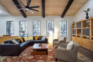 Una sala de estar moderna con un sofá seccional, una mesa de centro y una chimenea. Hay ventanas amplias con vistas a árboles.