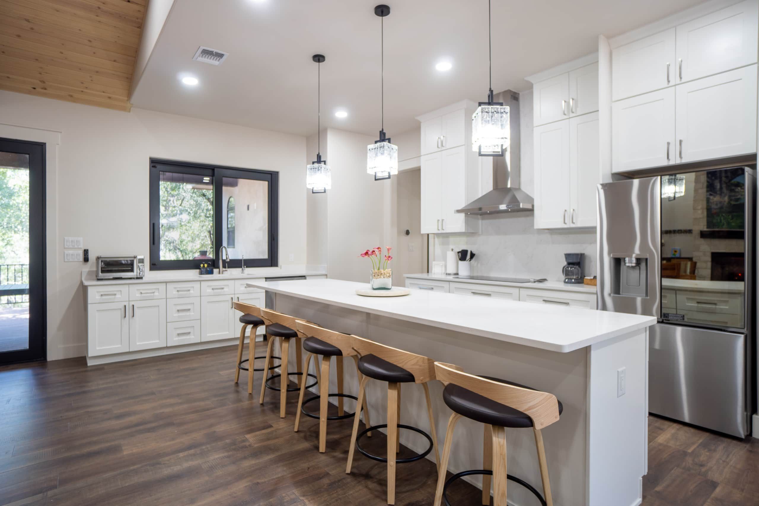 Una cocina moderna con armarios blancos, una isla grande con taburetes de bar, iluminación colgante y un refrigerador de acero inoxidable.
