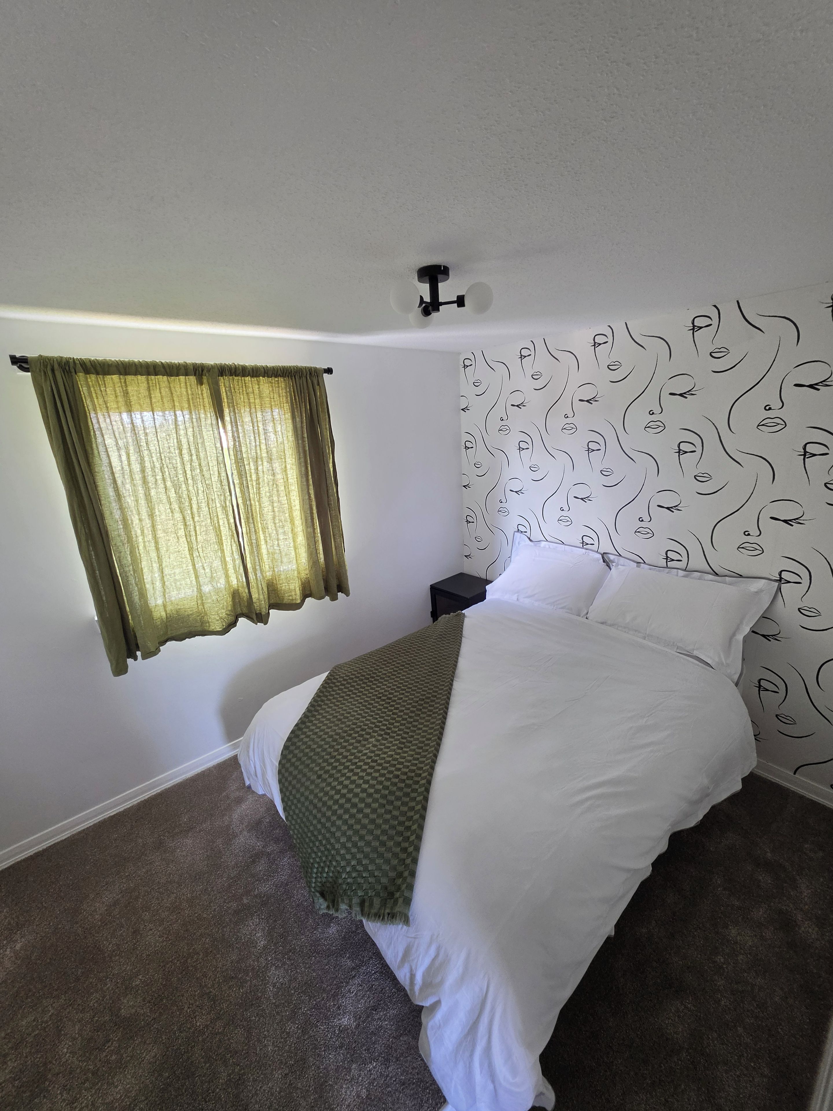 Un dormitorio con una cama, una ventana con cortinas, una pared con diseño de rostro y una mesita de noche pequeña.
