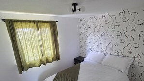 Un dormitorio con una cama, una ventana con cortinas, una pared con diseño de rostro y una mesita de noche pequeña.