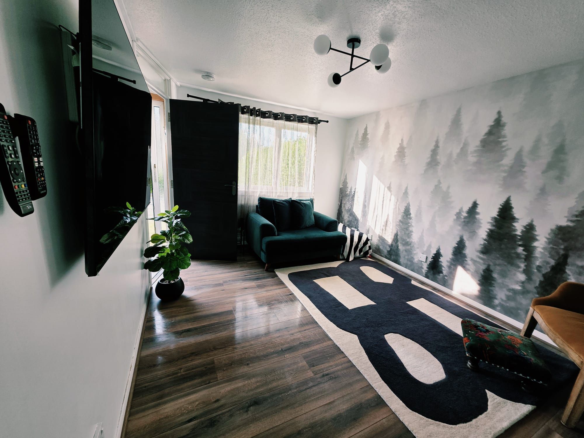 Una sala de estar moderna con un ventanal grande, un sofá verde, un tapete negro y un mural de un bosque en la pared.