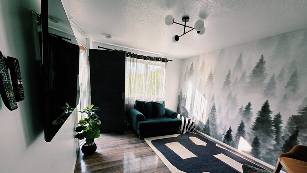 Una sala de estar moderna con un ventanal grande, un sofá verde, un tapete negro y un mural de un bosque en la pared.
