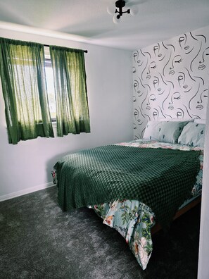 Un dormitorio con cama, una ventana con cortinas verdes, cabecera estampada y piso alfombrado.