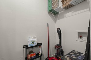 A corner of a room with a white wall, a black shelf holding cleaning supplies, a red broom, and a vacuum cleaner. - The Sundown - Freshly Renovated, Cozy North Loop Home w/ Hot Tub (Austin)
