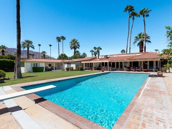 Ein Pool mit einer gefliesten Terrasse, umgeben von Palmen und einem Gebäude mit einem roten Ziegeldach.
