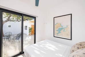 Une chambre avec un lit, une carte accrochée au mur et une porte-fenêtre coulissante donnant sur un espace extérieur aménagé avec des chaises et un arbre.