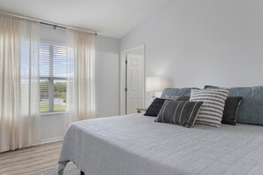 Une chambre à coucher avec un lit, des oreillers, une table de chevet, une lampe et une fenêtre avec des rideaux.