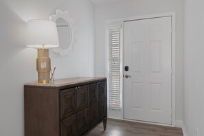 Un couloir avec une commode en bois, une lampe, une porte blanche et une fenêtre avec des stores.