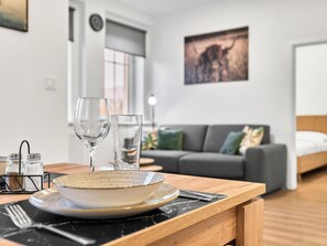Un comedor con una mesa de madera, dos copas de vino, un salero y pimentero, y un bol. Un sofá gris y una cama se ven al fondo.