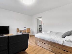 A modern hotel room with a flat-screen TV, a wooden bed, a grey sofa, a wooden dining table with chairs, and a kitchenette in the background. - Mánesova by Interhome (Jachymov)