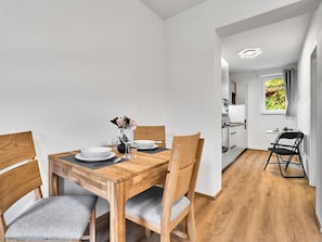 A modern dining area with a wooden table set for two, a kitchen in the background, and a view of greenery through the window. - Mánesova by Interhome (Jachymov)