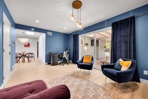 Ein modernes Wohnzimmer mit blauer Wand, einem dunkelblauen Sessel, einem roten Sofa und einem Essbereich mit Holzmöbeln.