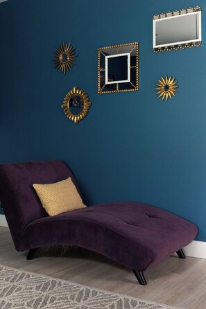 Ein violettes Sofa mit einem gemusterten Kissen vor einer blauen Wand, umgeben von dekorativen Spiegeln und Wandbildern im Sonnenstrahl-Design.
