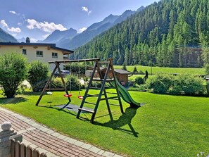 A playground with swings and a slide, surrounded by a well-maintained lawn, a building, and a mountain backdrop. - Ferienhaus Brigitte by Interhome (Längenfeld)
