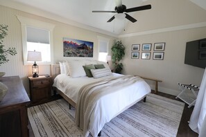 Une chambre à coucher comprenant un lit, des tables de chevet, un ventilateur de plafond, une fenêtre avec des stores, un tableau, une plante en pot et un téléviseur.