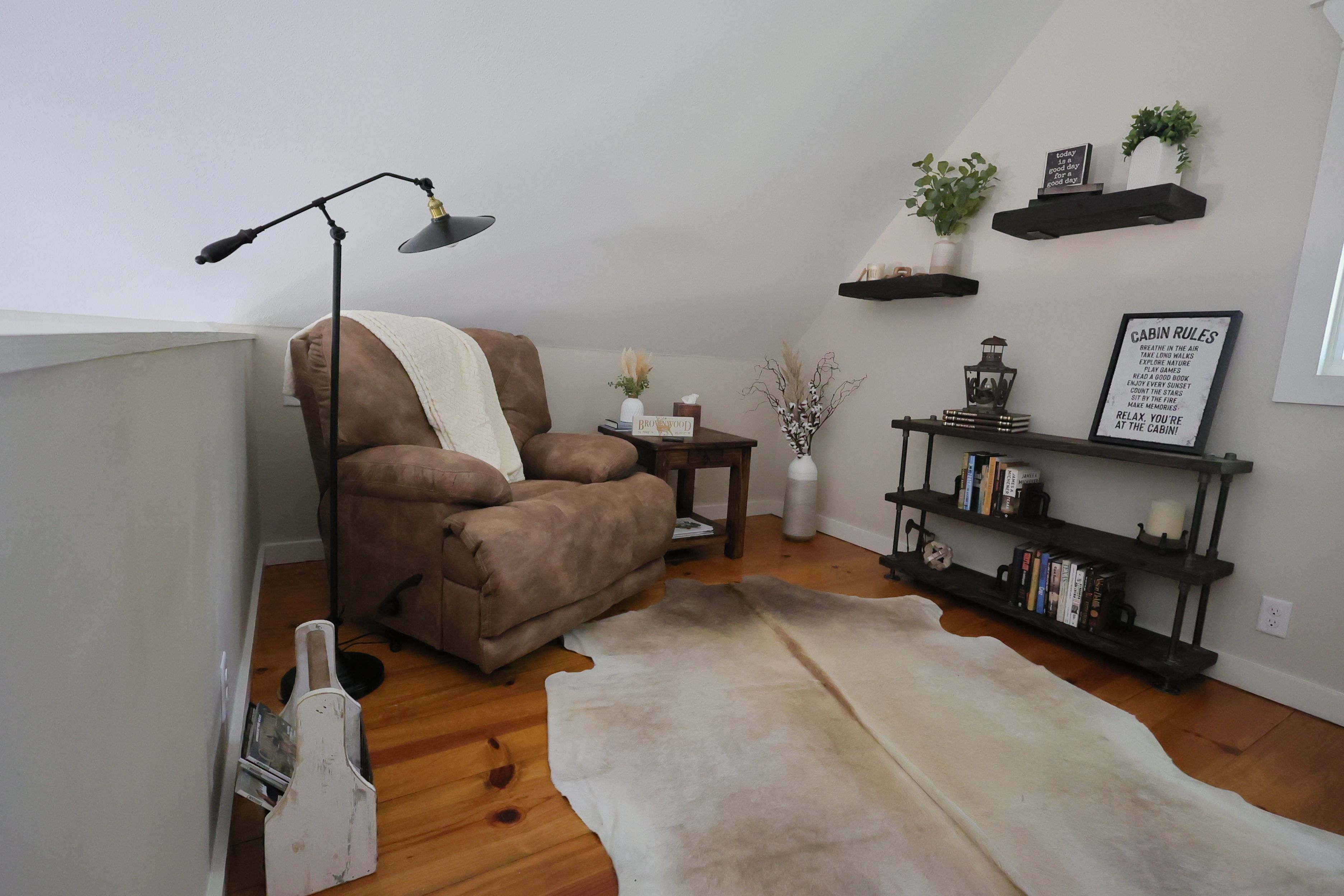 A cozy corner with a brown armchair, a floor lamp, a white rug, a bookshelf with books, and decorative items on shelves.