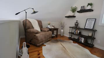 A cozy corner with a brown armchair, a floor lamp, a white rug, a bookshelf with books, and decorative items on shelves.