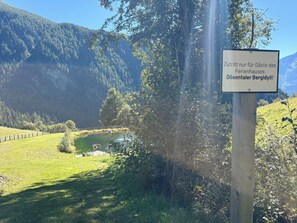 Un cartel con texto en idioma extranjero, un cuerpo de agua, una zona cubierta de hierba y montañas boscosas al fondo.