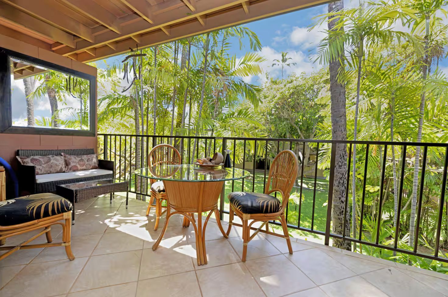 A balcony with a glass table and chairs, a sofa, and a view of tropical trees.