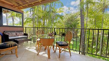 A balcony with a glass table and chairs, a sofa, and a view of tropical trees.
