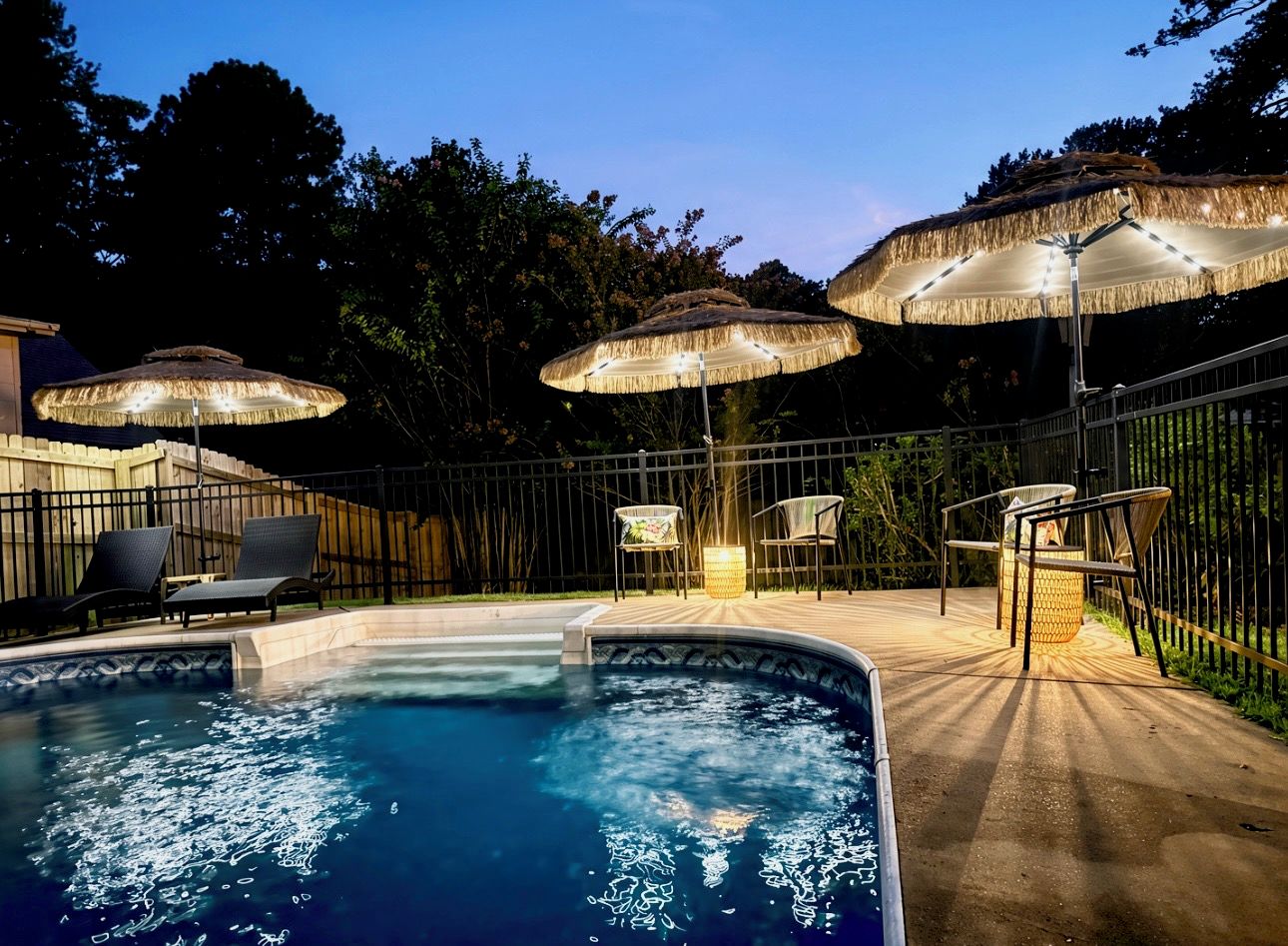 A swimming pool with lounge chairs and umbrellas, surrounded by a fence and trees.