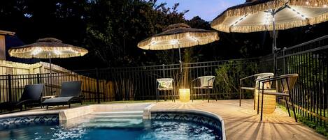 A swimming pool with lounge chairs and umbrellas, surrounded by a fence and trees.