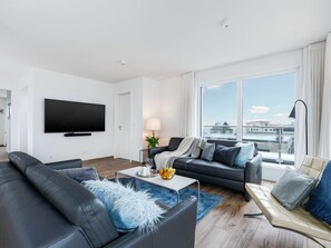 Ein modernes Wohnzimmer mit einem Flachbildfernseher, zwei Sofas, einem Couchtisch und Blick auf die Stadt durch große Fenster.